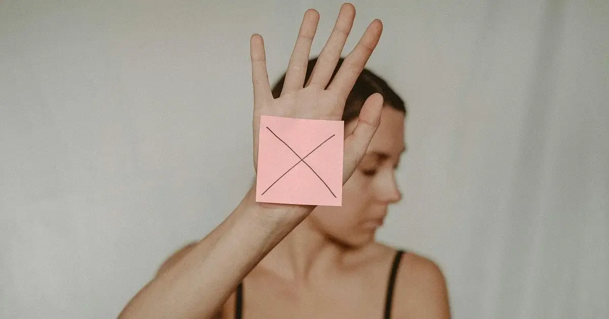 A girl holding up her hand with a sticky note with an x on, symbolizing 'no'