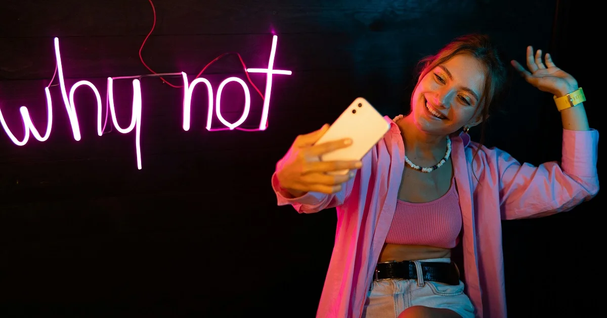 Woman taking selfie while expressing a why-not kind of attitute with a neon-sign of the expression in the background.